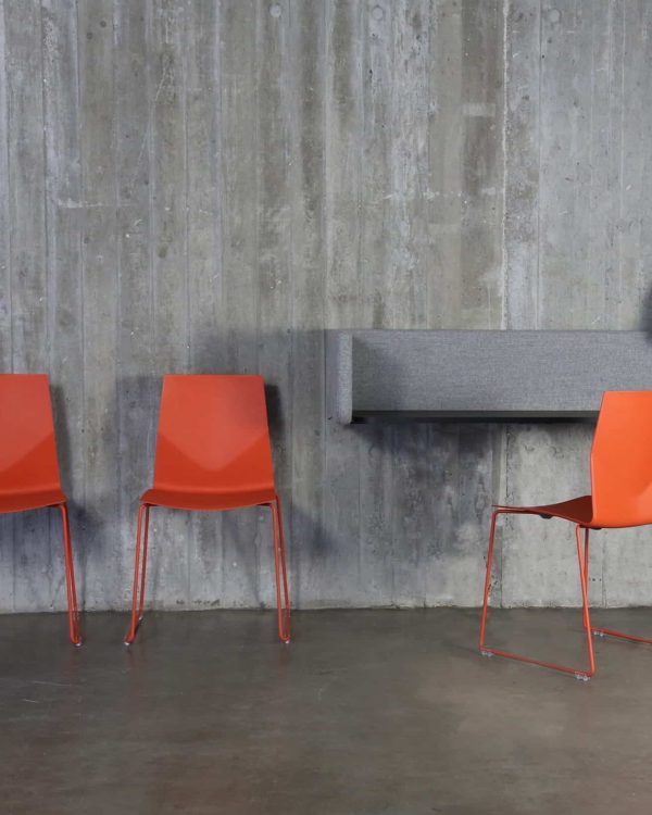 Three orange chairs in front of a desk workstation and concrete wall.