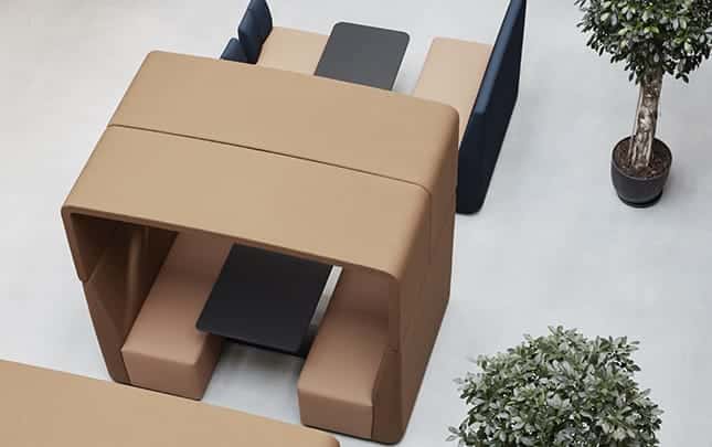 A group of office work booths, chairs and tables in a room with a tree in the background.