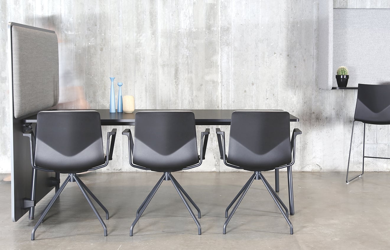 A black table with office screen divider and chairs in a room with a concrete wall.