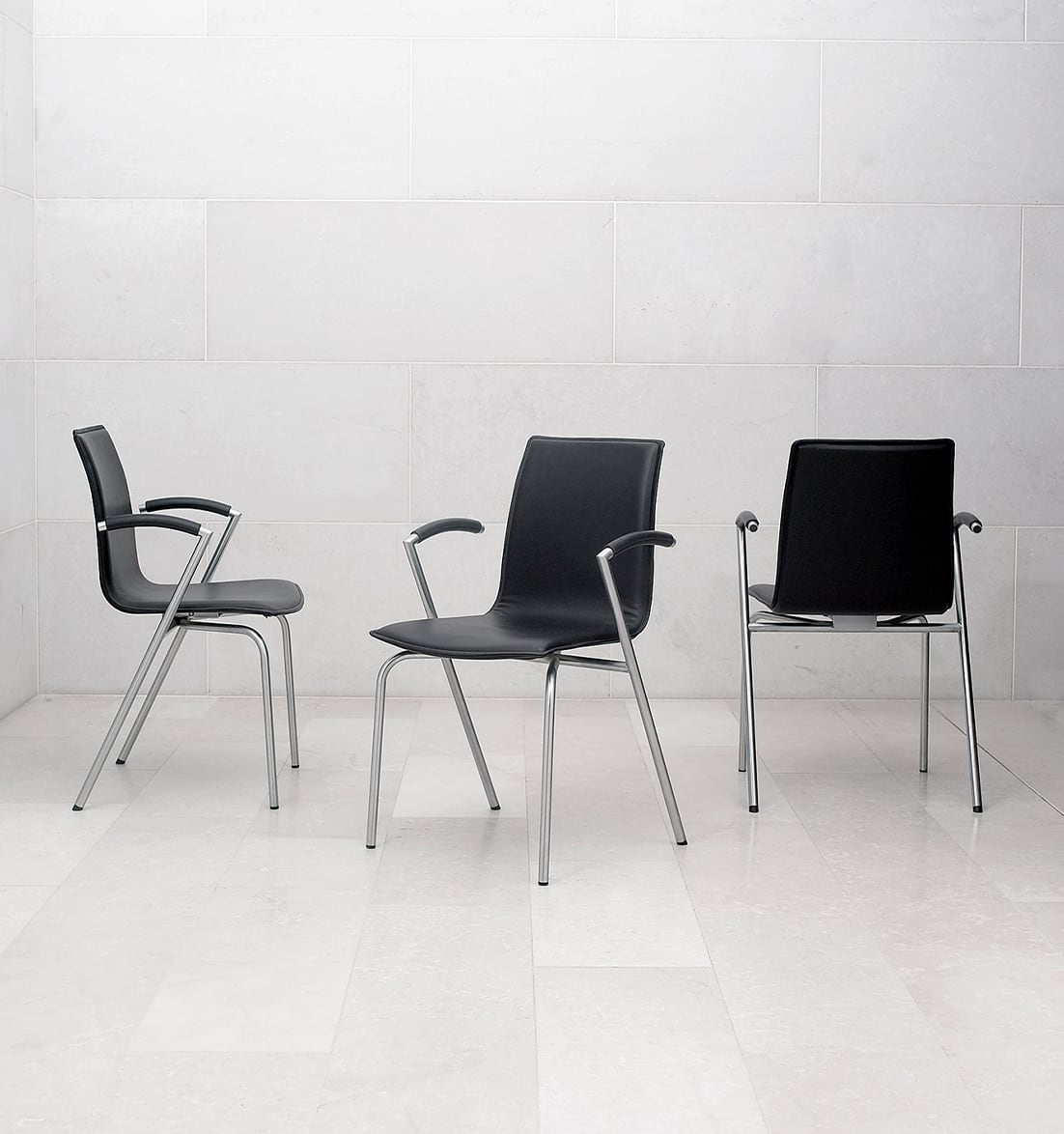 Three black chairs with arm rests in front of a white wall.