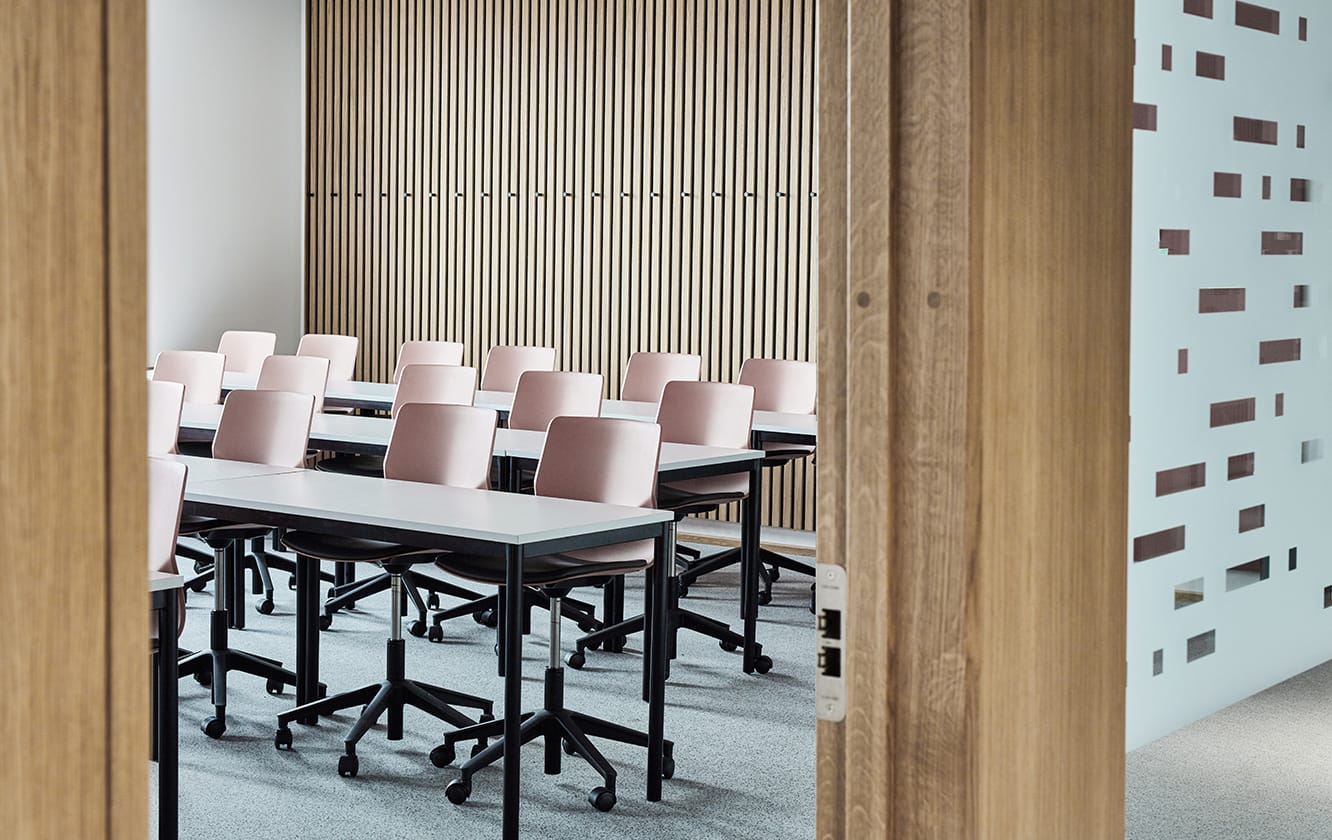 A conference room with pink office desk chairs behind office desks and wooden walls.