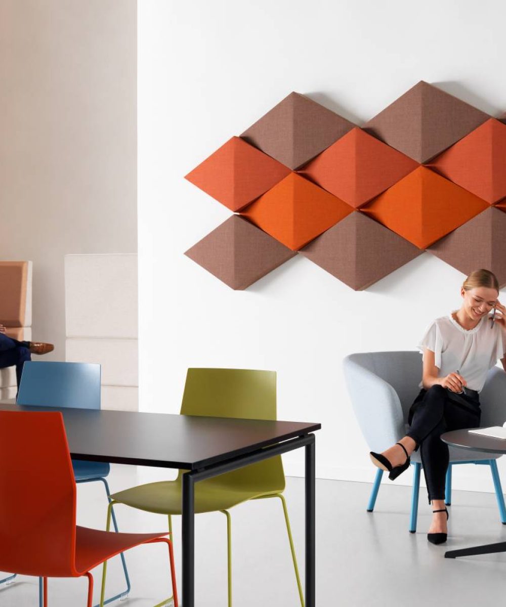 A woman sits at a table in an office with colourful acoustic panels for offices behind her. A man sits in an office work booth. Office furniture is spread around the office.