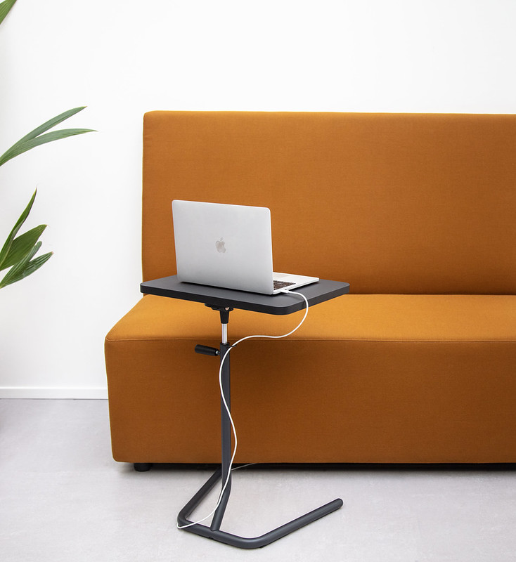 A laptop is sitting on a Y-table adjustable height work table next to a plant and office sofa.
