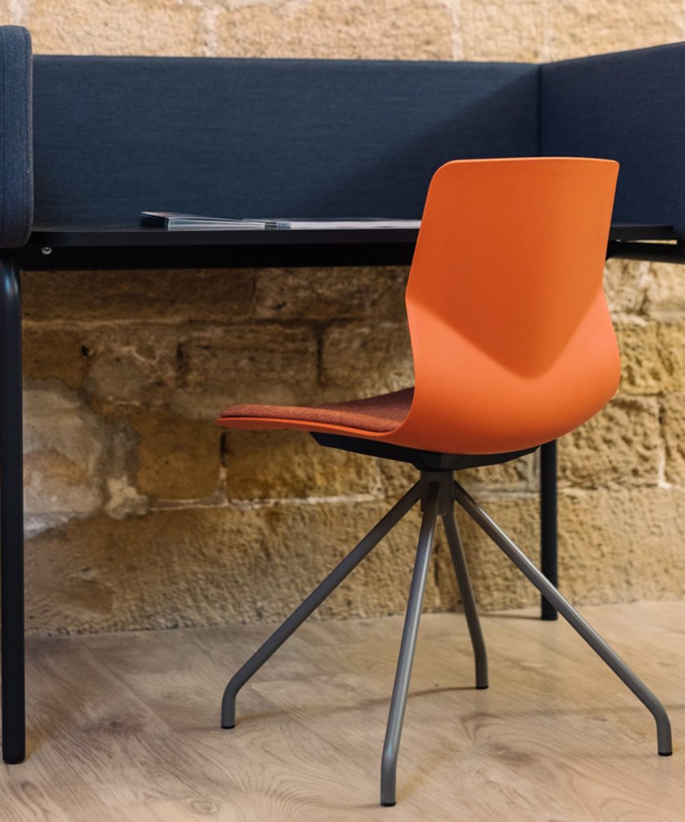 A desk with an orange chair and a work deskstation with a wall behind it.